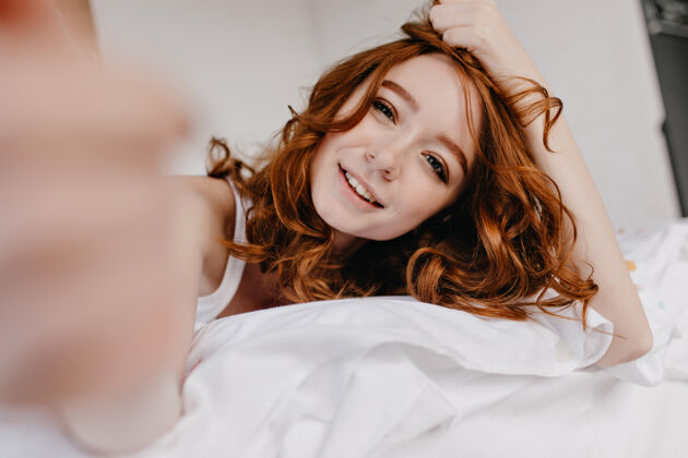 卷发美丽的姜黄色头发的白色女孩在早晨放松可爱的白人年轻女士在床上自拍的室内照片肖像青春姜