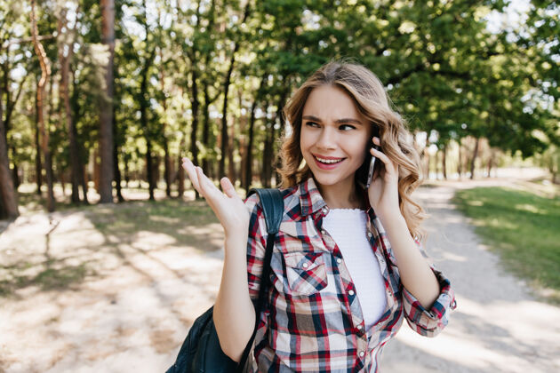 休息在公园散步的时候 心情很好的漂亮女孩打电话给朋友一个可爱的卷发女人拿着背包和电话摆姿势的户外镜头快乐活跃旅行者