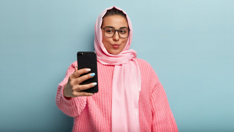 高兴美丽的黑发女士录制视频 嘴唇合拢 自拍 捕捉新的面貌 在粉红色围巾和毛衣的姿势 张贴照片在网上的追随者 使室内凉爽的镜头智能手机小玩意毛衣