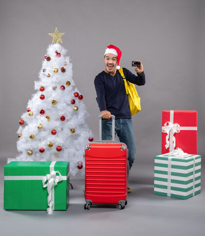 圣诞树带着圣诞帽的兴奋的年轻人拿着一个红色的手提箱 上面有一张灰色的卡片兴奋的年轻人红色圣诞帽