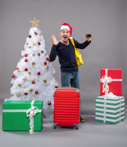 圣诞带着卡片和黄色背包的年轻人站在圣诞树旁欢欣鼓舞 礼物是灰色的快乐的圣诞帽圣诞老人