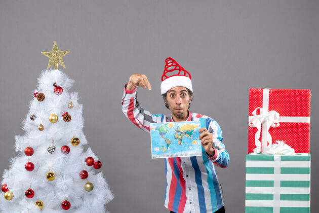 圣诞树前视图惊讶的男子与螺旋弹簧圣诞帽指向世界地图目的地快乐盒子