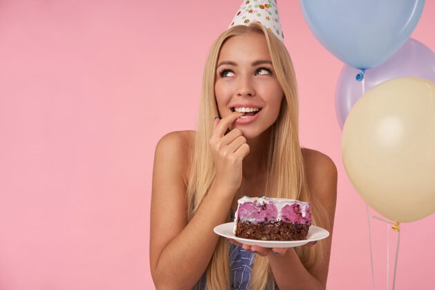 摆姿势快乐的美女在五彩缤纷的气球里欢呼雀跃 用美味的蛋糕庆祝生日 积极地看着身边 憧憬着未来 在粉色的背景下与世隔绝空气年轻饮食