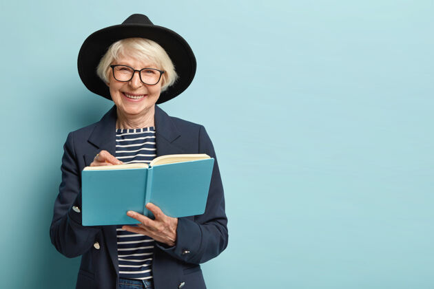 女人高兴的女性退休者画像在日记里写计划策略 有着漂亮的聪明外表 戴着眼镜和黑帽子 隔着空旷的蓝色墙壁女商人用笔记本退休皱纹头饰