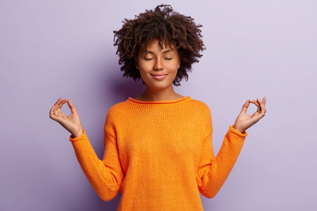 衣服容貌端庄的女人在室内冥想 手举着mudra的手势 有迷人的微笑 闭着眼睛 穿着橙色的衣服 模特翻过紫色的墙壁手势冥想的概念非洲手势个人