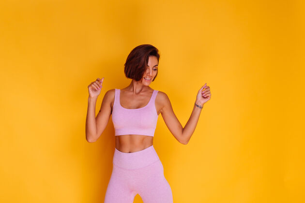 腹肌体育女站在黄色的墙上 展示她的腹肌 满意的健身训练和饮食的结果 有一个愉快的面部表情 穿着运动上衣和紧身裤形状锻炼健康