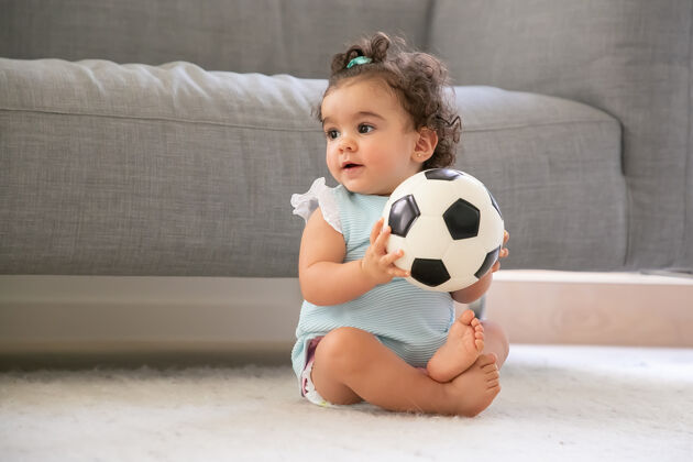 赛车正面黑发女婴穿着淡蓝色的衣服坐在家里的地板上 看着别处 踢足球复制空间孩子在家和童年的概念卷发婴儿年轻