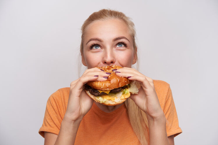 休闲可爱的金发女郎的肖像 随意的发型 食欲旺盛地吃着新鲜的汉堡包 愉快地向上看 在白色背景上摆姿势垃圾表情持有