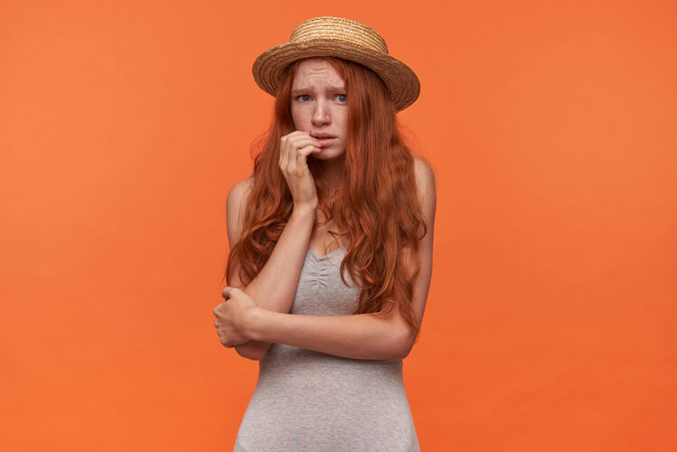 折叠一头卷发的年轻红发女子 穿着灰色衬衫 头戴草帽 手放在脸上 面对镜头 她显得很害怕 在橙色背景下摆姿势手害怕肖像
