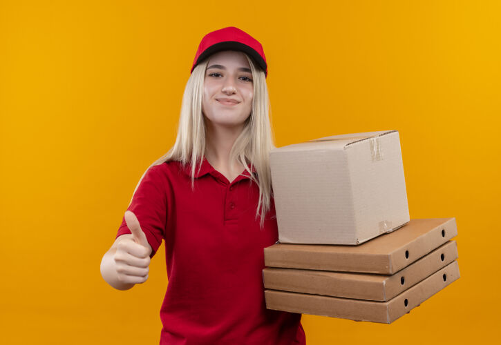 送货一个穿着红色t恤 戴着帽子 拿着盒子的年轻女孩 在孤立的橙色背景上竖起大拇指帽子高兴红色