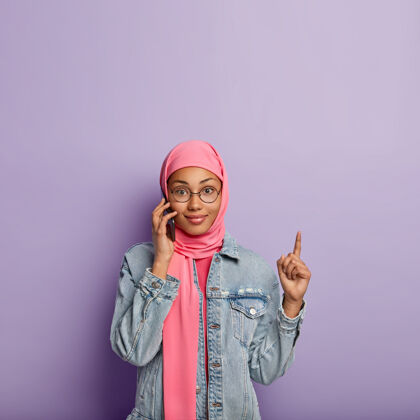 服装漂亮的女士和接线员谈论关税 手持现代手机 用食指指着上面的空白处 戴着粉色围巾 隔着紫色的墙电话指向手机