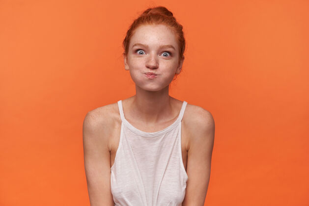 20多岁一张室内照片 一位年轻有趣的女士扎着红发 看着相机 鼓起脸颊 在白色上衣的橙色背景上做鬼脸圆形自然情绪