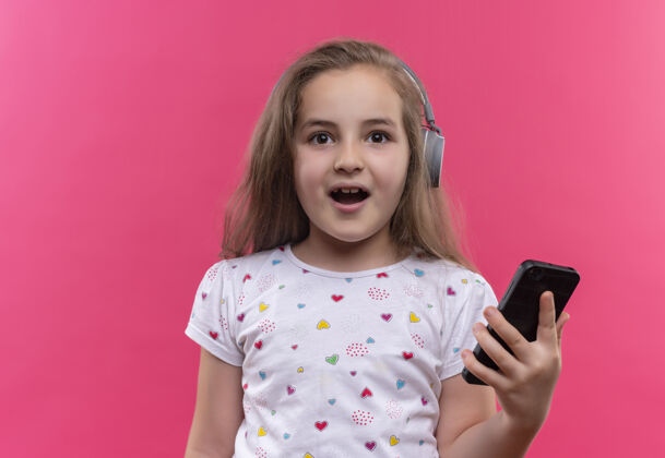 T恤微笑的小女孩穿着白色t恤 戴着耳机 拿着手机 背景是孤立的粉色粉色学校耳机