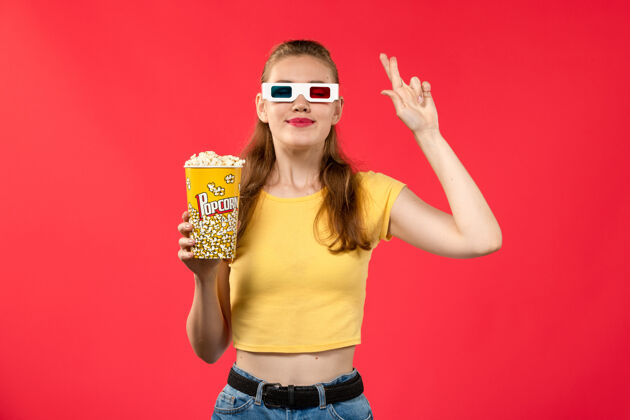 浅红色正面图年轻女性手持爆米花包 戴着d型太阳镜 站在浅红的墙上看电影电影院视图护目镜