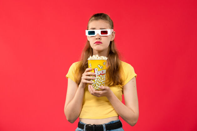 肖像正面图年轻女性在电影院拿着爆米花戴着d太阳镜在红墙电影院看电影女性的颜色电影院女性漂亮