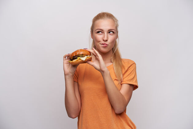 长摄影棚拍摄了一个积极的年轻漂亮的长发金发女性 一边吃着新鲜的汉堡 一边怀疑地看着一边 一边穿着橙色t恤 一边站在白色背景上不健康发型女性