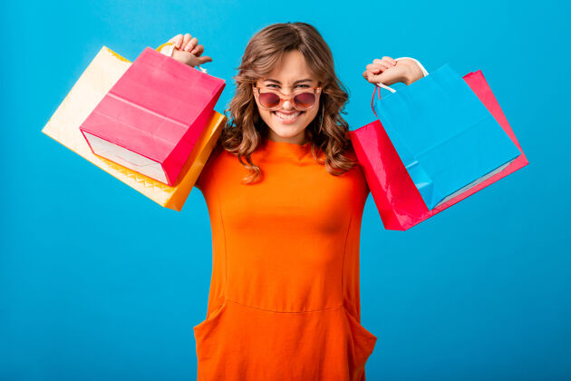 购买图片中的兴奋迷人的微笑时尚女性购物狂穿着橙色的新潮连衣裙拿着购物袋在蓝色的工作室背景隔离黑色开朗女士