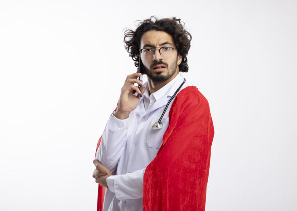 斗篷自信的年轻白种人超级英雄戴着眼镜 身穿医生制服 披着红色斗篷 脖子上戴着听诊器 站在一旁打电话穿着站着光学