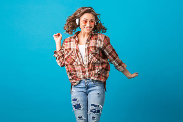 一穿着格子衬衫和牛仔裤 在蓝色摄影棚背景下 戴着粉色太阳镜 迷人的情绪化女人跳跃着滑稽疯狂的表情微笑肖像跳跃