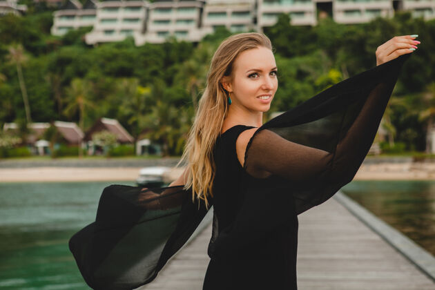 岛屿穿着黑色连衣裙的美丽迷人的女人在豪华度假酒店的码头上摆姿势 暑假 热带海滩女人身体赤脚