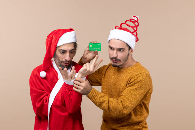 男性前视图两个困惑的家伙一个穿着圣诞外套 另一个拿着卡在米色孤立的背景上微笑微笑两个困惑的家伙