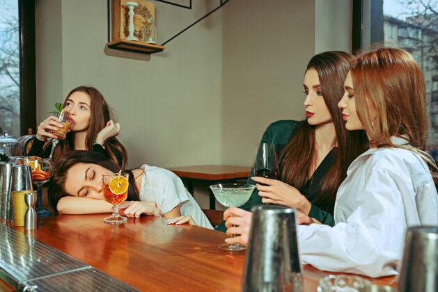 水果女朋友在酒吧喝酒她们坐在一张摆着鸡尾酒的木桌旁她们穿着休闲服杯子手团体