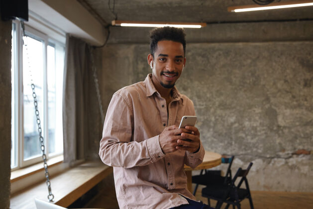 工作一个穿着米色衬衫的年轻黑皮肤男人在咖啡馆里摆姿势 举着智能手机 带着卡门式的微笑看着相机开朗胡须休闲