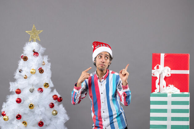 年轻人前视图：年轻人用手指指着站在白色圣诞树旁的东西人快乐圣诞节