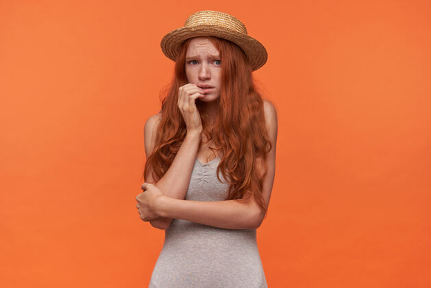 折叠一头卷发的年轻红发女子 穿着灰色衬衫 头戴草帽 手放在脸上 面对镜头 她显得很害怕 在橙色背景下摆姿势手害怕肖像