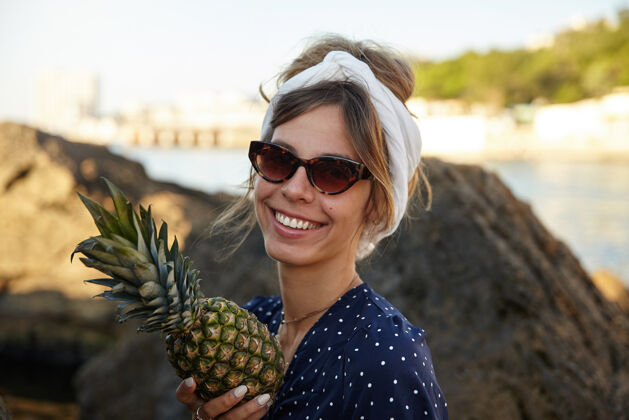 宽美丽的年轻黑发女子 随意的发型 戴着头巾 面带幸福的笑容 在阳光明媚的夏日 手里拿着新鲜的菠萝 在海湾上空摆着姿势女人休闲微笑