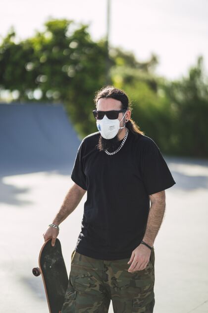 滑板一个戴着口罩的大胡子男人拿着滑板在公园散步的垂直镜头面罩病毒太阳