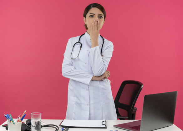 电脑惊讶的年轻女医生穿着医用长袍 听诊器站在办公桌后面 用医疗工具在电脑上工作 嘴上盖着复印空间医疗罩子惊讶