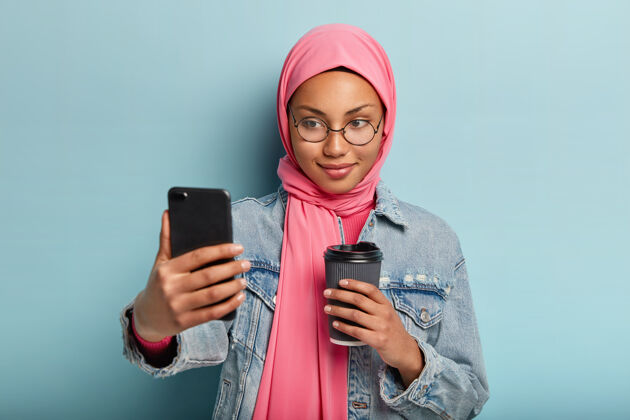 站立一个看起来很愉快的女孩喝外卖咖啡 自拍肖像或视频通话 穿着时髦的牛仔夹克和头巾 在社交网络中与追随者分享图片眼镜智能手机女人