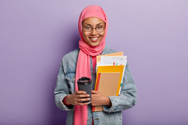 围巾快乐的伊斯兰女人的孤立镜头拿着外卖咖啡 拿着带着文件的记事本 有时间休息和热饮 戴着传统的粉色头巾 表达良好的情绪 孤立在紫色上咖啡休息传统学习