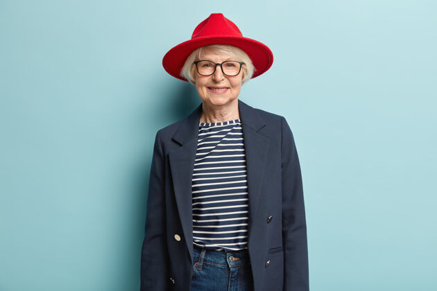 祖母时尚的白发女士 满脸皱纹 戴着红色时髦的帽子 夹克和牛仔裤 微笑愉快年龄时尚乐观