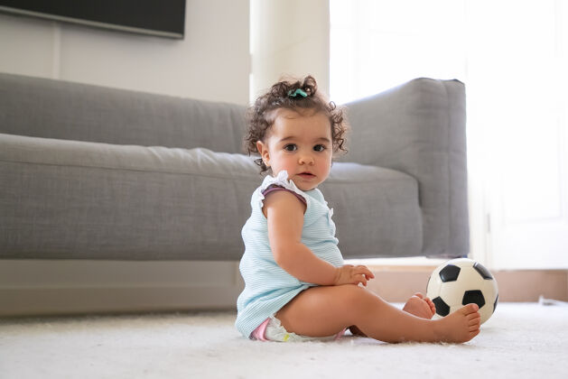 沙发甜美严肃的黑发女婴穿着淡蓝色的衣服坐在地板上 手里拿着足球 a.侧视图孩子在家和童年的概念侧视图可爱侧
