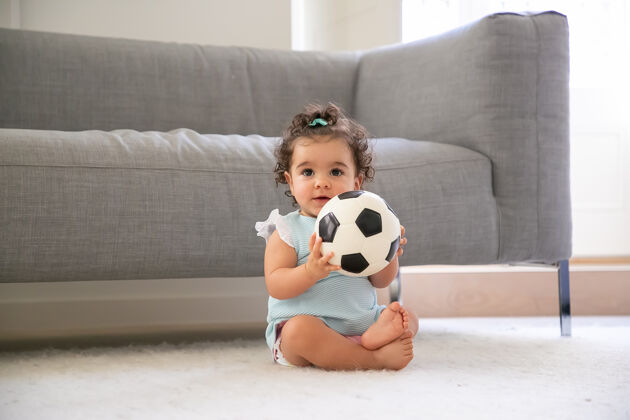 赛车快乐的黑色卷发女婴 穿着淡蓝色的衣服 坐在家里的地板上 看着别处 踢足球正面视图孩子在家和童年的概念正面弓混合