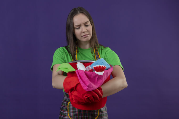 戴着悲伤的清洁年轻女孩穿着制服 戴着红色手套 手持清洁工具 背景是孤立的紫色工具手套清洁