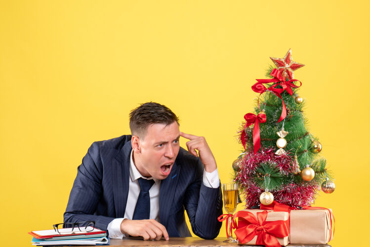 男人年轻人边喊边坐在圣诞树旁的桌子上 黄色的礼物上礼物年轻专业