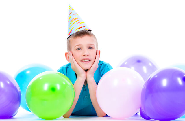 躺着穿着蓝色t恤的快乐微笑的男孩躺在地板上 手里拿着五颜六色的气球 竖起大拇指——孤立地站在白色的地板上孩子玩气球
