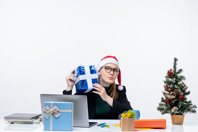 坐着圣诞气氛与戴着圣诞老人帽子和眼镜的年轻女子坐在一张桌子上 拿着她的礼物令人惊讶的白色背景办公室圣诞老人礼物