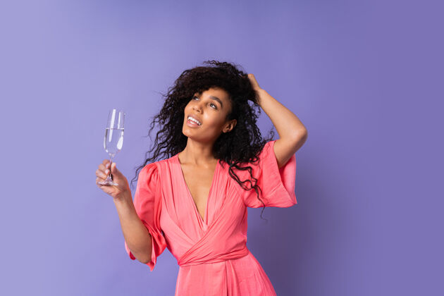 摆姿势一位年轻快乐的巴西女性 卷发 穿着粉色时髦的裙子 拿着一杯咖啡摆姿势紫色墙壁上的香槟派对气氛紫罗兰混合时尚
