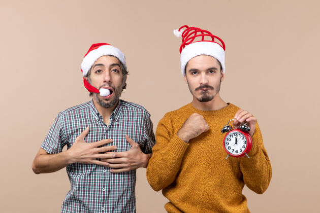 男人前视图两个圣诞老人一个把手放在胸前 另一个拿着闹钟 背景是米色的圣诞节帽子时钟