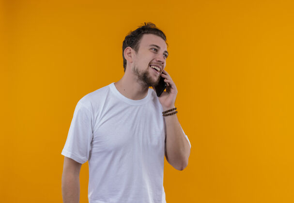 T恤微笑的年轻人穿着白色t恤在孤立的橙色背景下讲电话橙色男人穿着