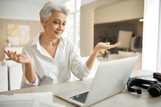 办公室情绪成熟的女员工穿着白衬衫在家工作 坐在桌边拿着笔记本电脑 做着无助的手势 耸耸肩 虚拟在线聊天办公桌通信技术