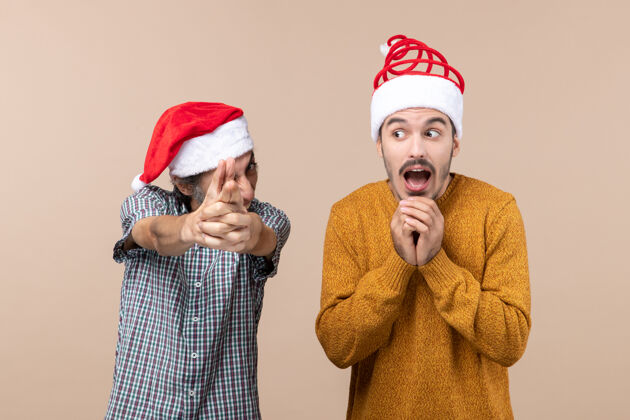 搞笑正面图两个戴着圣诞帽的滑稽家伙 一个在做手指枪的手势 另一个在米色孤立的背景上害怕害怕帽子两个