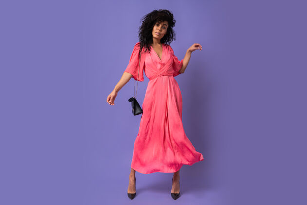 蓝紫色身着粉色派对礼服的优雅黑人女子在紫色墙壁上玩得很开心穿高跟鞋全长度肖像非洲头发