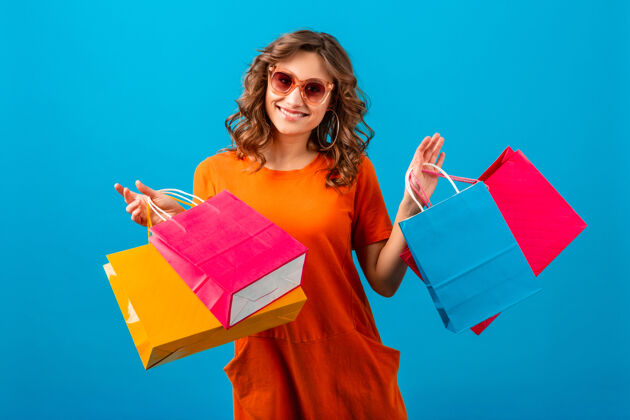 购物者迷人的快乐情绪微笑时尚的女人购物狂在橙色时尚的超大礼服举行购物袋蓝色工作室背景隔离蓝色销售太阳镜