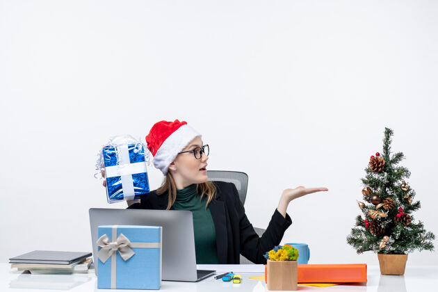 眼镜圣诞气氛：戴着圣诞老人帽戴着眼镜的年轻女子坐在桌子旁 手里拿着礼物 在白色背景下问问题圣诞老人举行年轻女子