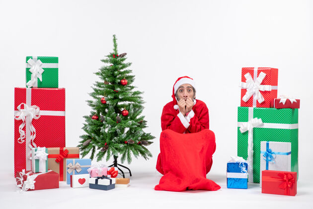 微笑吓坏了的年轻人打扮成圣诞老人 拿着礼物和装饰好的圣诞树坐在白色背景的地上礼物装饰坐着